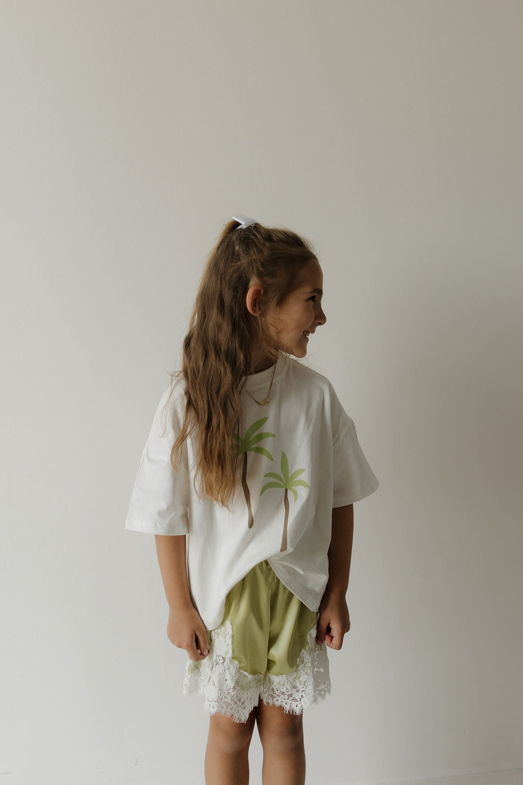 A young girl with long brown hair in a ponytail wears the forever french baby Child Cotton Tee | Palm, featuring palm trees, paired with green lace-trimmed shorts. She stands smiling and looking to the side against a plain white wall.