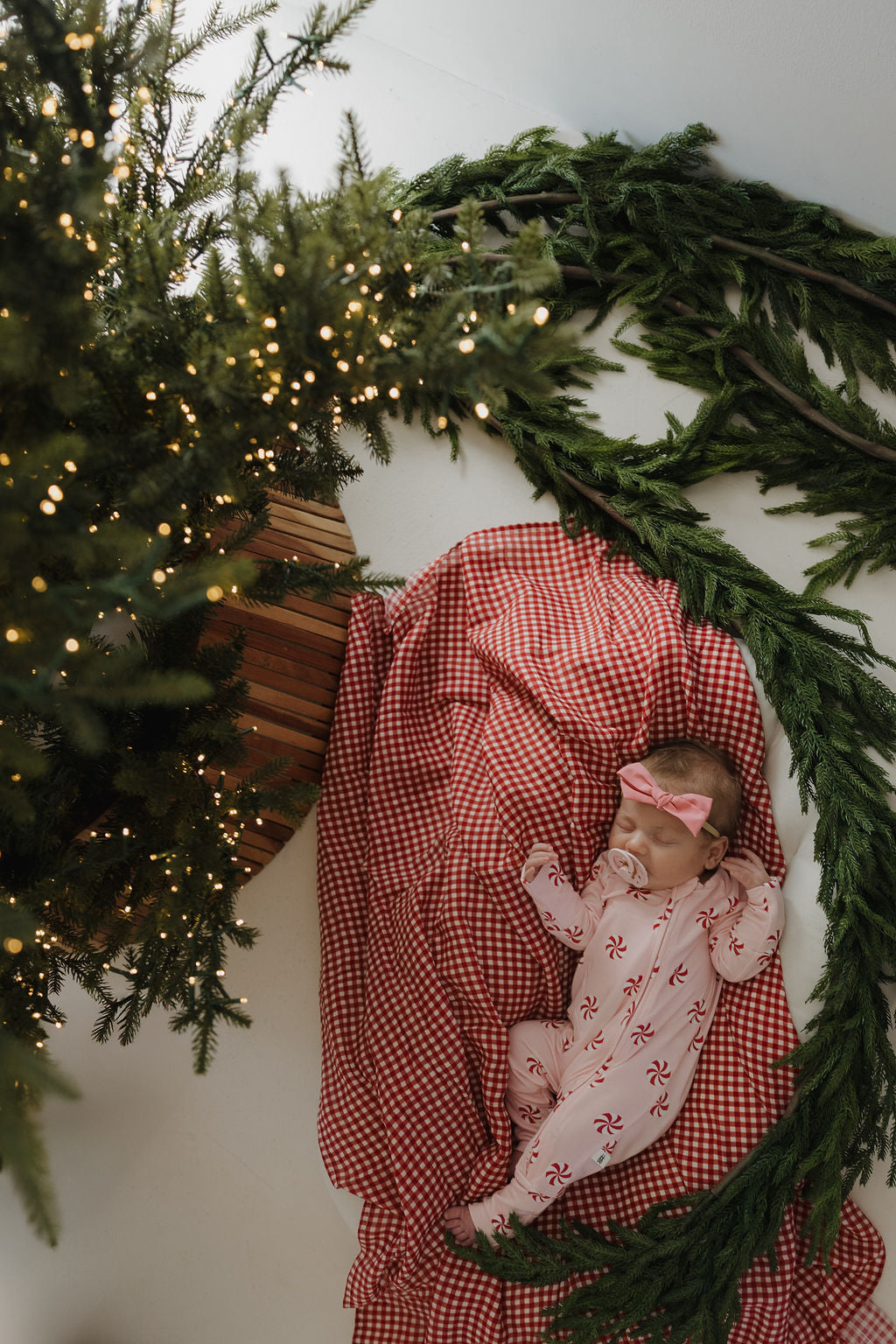 A baby in festive pajamas and a pink bow headband naps on the forever french baby Muslin Swaddle | Red Gingham, surrounded by a decorated Christmas tree and green garland, viewed from above.