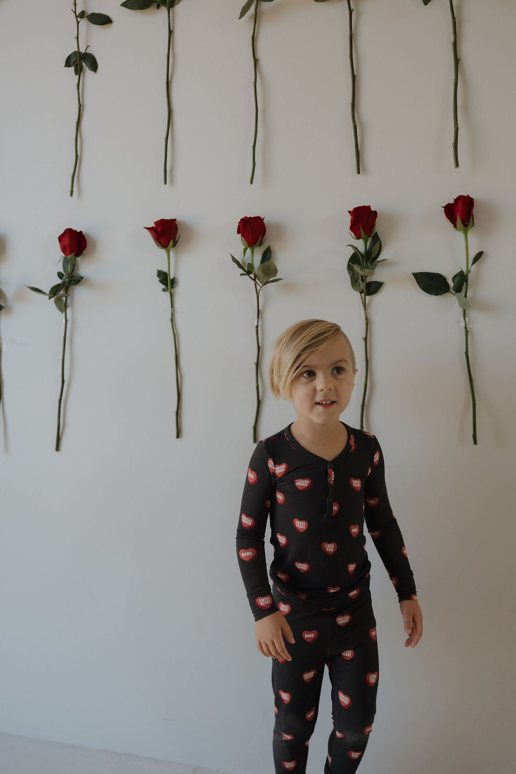 A young child with blond hair stands by a white wall of red roses, wearing forever french baby's Bamboo Two Piece Pajamas | Love Notes – Charcoal—hypoallergenic black kids sleepwear with a playful red lip print.