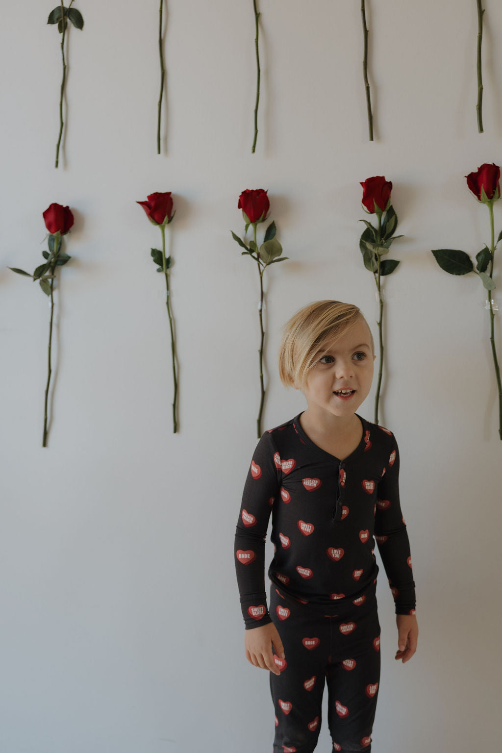 A young child wears forever french baby's Bamboo Two Piece Pajamas | Love Notes – Charcoal, featuring a red lips print, standing before a white wall adorned with vertical rows of red roses—showcasing soft, breathable bamboo comfort.