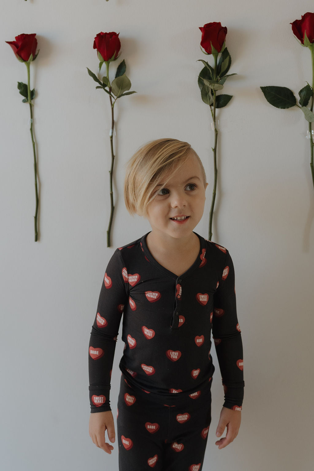 A young blonde child wears forever french baby Bamboo Two Piece Pajamas | Love Notes – Charcoal, featuring red lips, while standing in front of a white wall adorned with vertical rows of red roses.