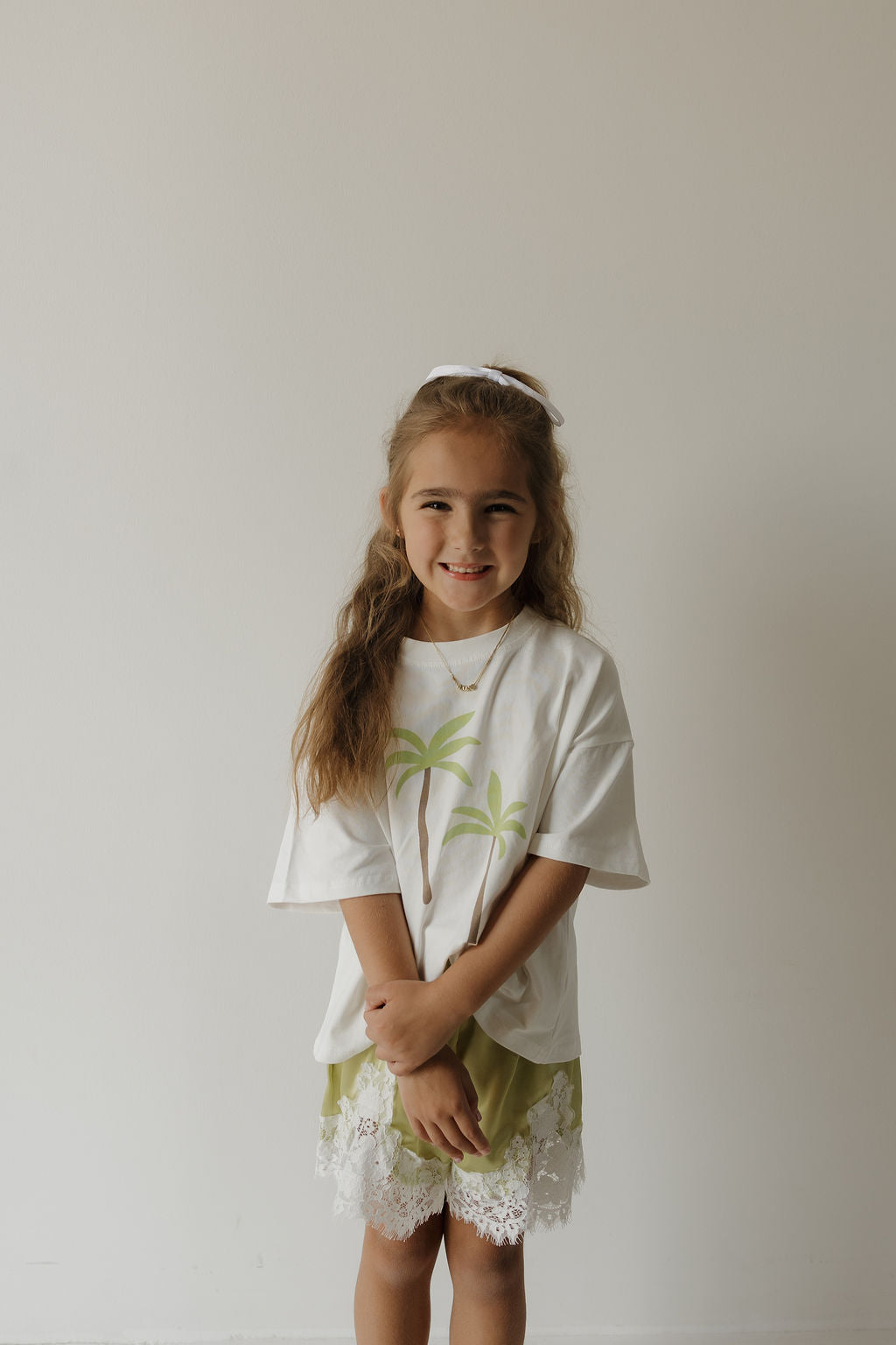 A young girl with long hair wears a white t-shirt with palm tree prints, a white hairband, and forever french baby’s Girl’s Satin Shorts in the Prickly Pear color, smiling as she stands against a plain light-colored wall.