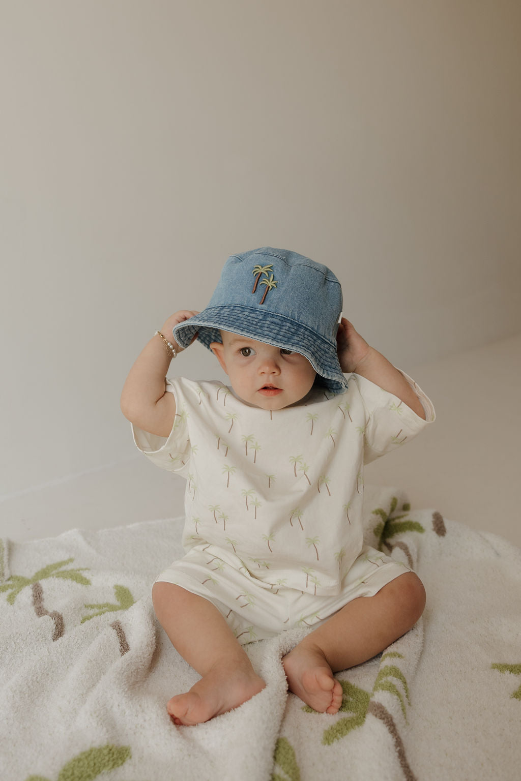A baby sits on a blanket, wearing the forever french baby “Child Short Set | Palm”—a cream cotton outfit featuring a subtle palm tree print—paired with a blue bucket hat. The baby is holding the brim of the hat and gazing curiously at the camera.
