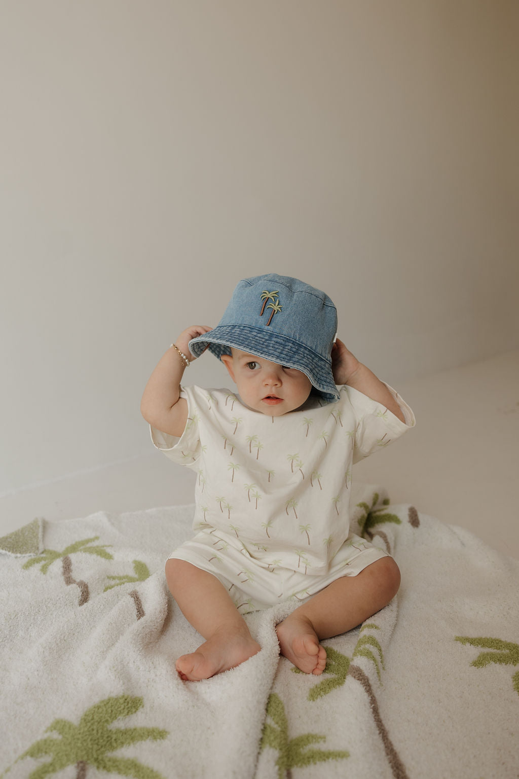 A baby wearing a light-colored outfit and the forever french baby Bucket Hat | Palm sits on a blanket with green palm tree designs, looking at the camera while holding the brim of the hat.