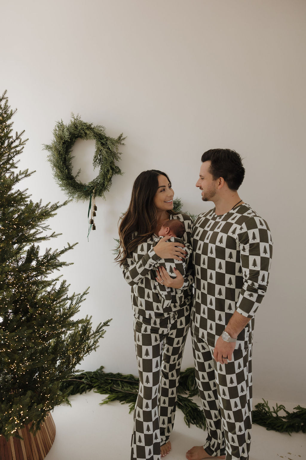 A happy couple in forever french baby Men's Bamboo Pajamas | Christmas Tree Checkerboard pose barefoot with their baby by a festive tree and wreath, celebrating the season in comfy, hypo-allergenic bamboo clothing.