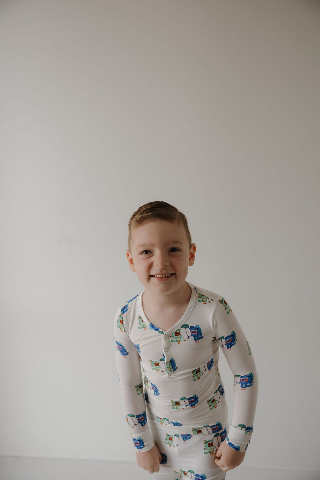 A young boy with short light brown hair smiles at the camera wearing forever french baby’s Bamboo Two Piece Pajamas | Charli's Monsters—white hypoallergenic kidswear with colorful cartoon prints—standing against a plain white background.