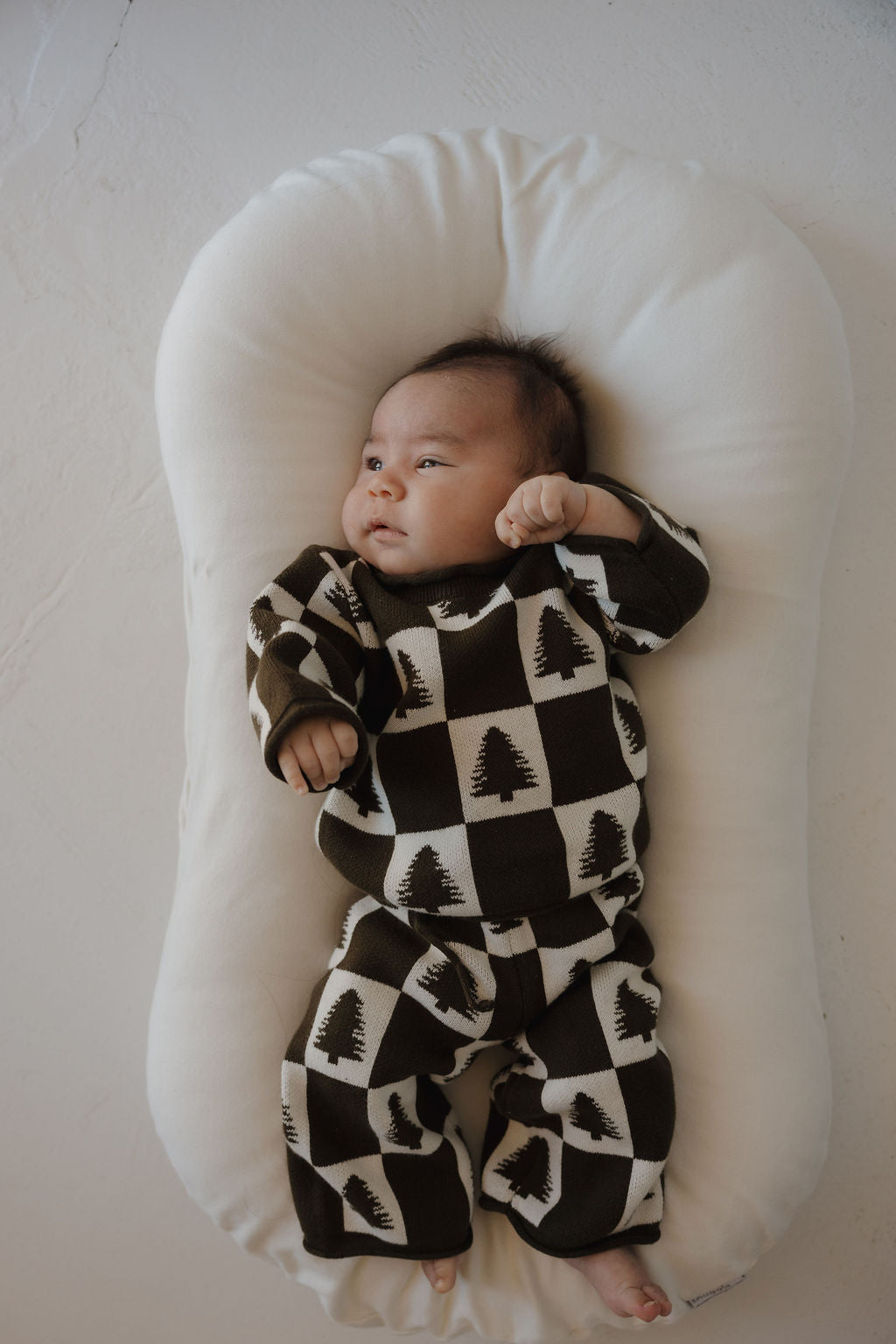 A baby in the forever french baby Knit Pant Set | Christmas Tree Checkerboard—made from 100% cotton—rests on a soft white cushion, gazing off to the side with one arm raised. The checkered tree design makes it perfect for any winter wardrobe.