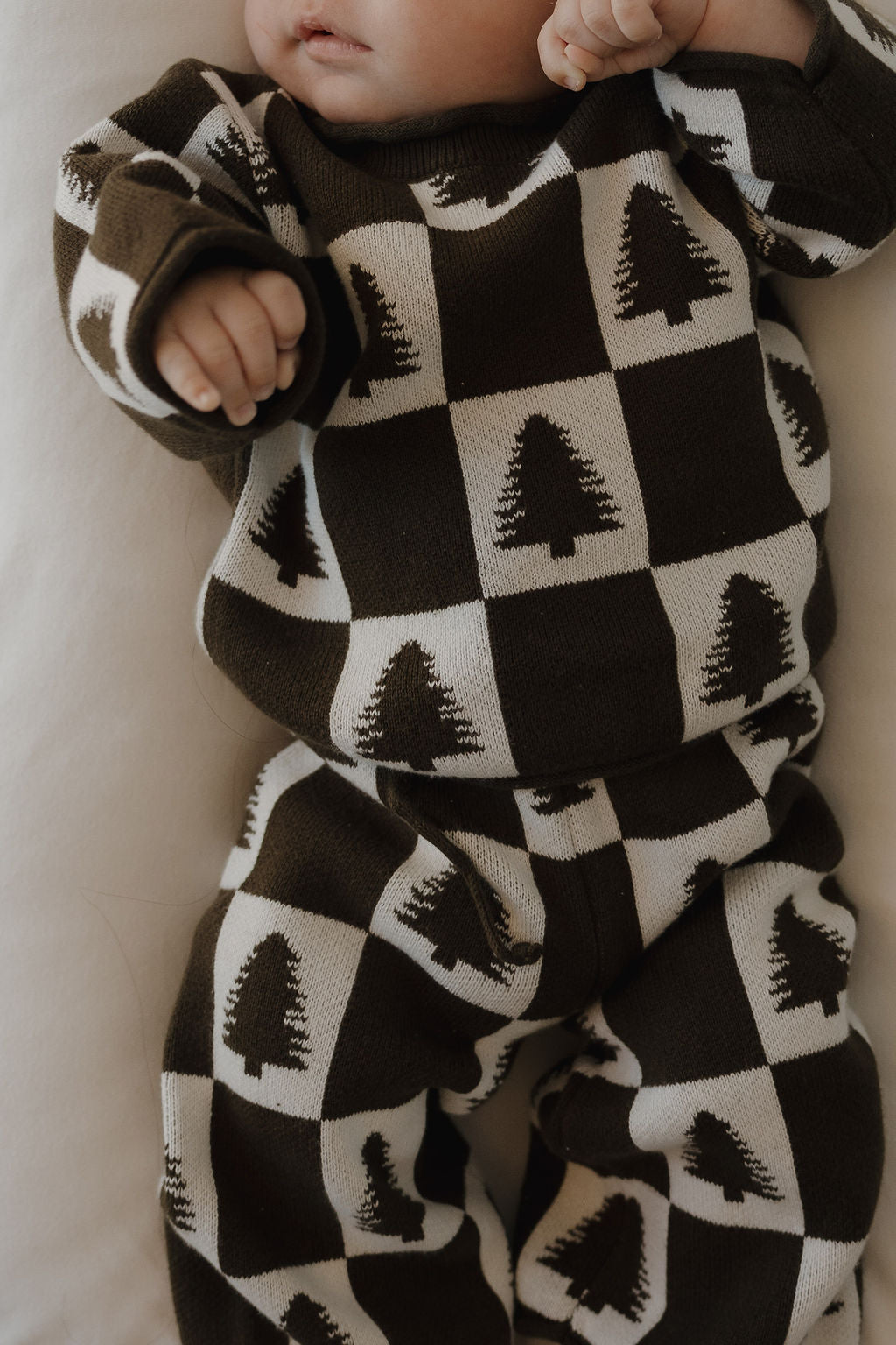 A baby in the forever french baby Knit Pant Set | Christmas Tree Checkerboard, featuring a brown and white checkerboard with pine tree designs, is ready for winter while lying on a light-colored surface.