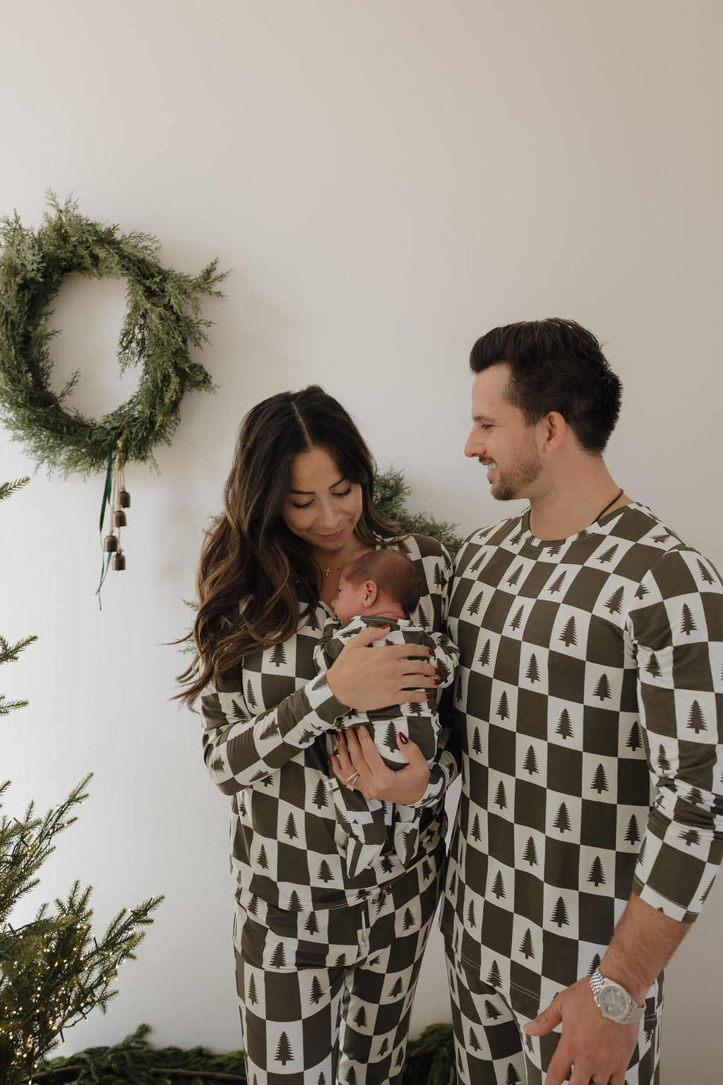 A couple wears matching forever french baby Bamboo Zip Pajamas in Christmas Tree Checkerboard print, while the woman cradles a baby in breathable, hypo-allergenic pajamas. A festive wreath and greenery decorate the background.