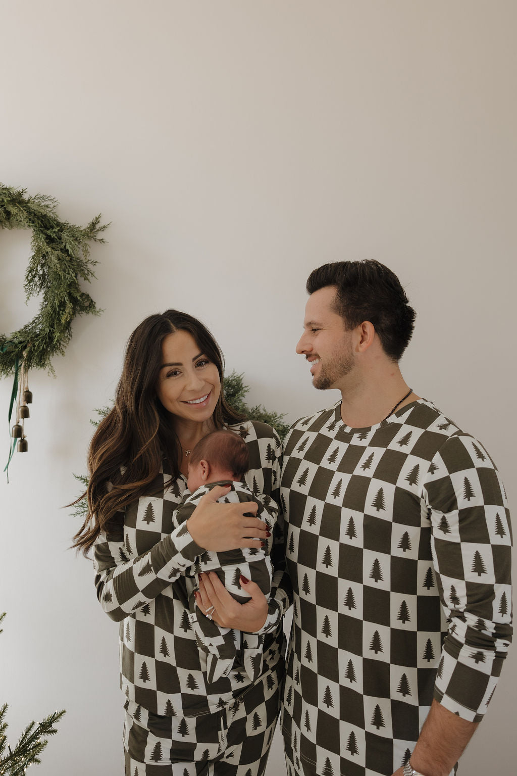 A smiling couple wears matching forever french baby Men's Bamboo Pajamas in Christmas Tree Checkerboard print; the woman holds a newborn in similar hypo-allergenic bamboo clothing, with festive greenery in the background.