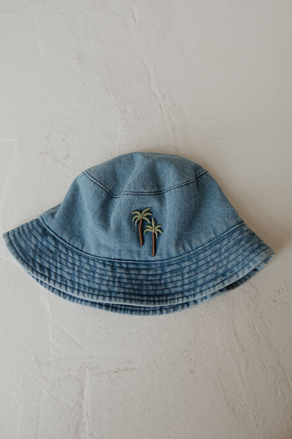 A light blue denim “Bucket Hat | Palm” from forever french baby, featuring two small embroidered palm trees on the front, is displayed on a textured white surface.