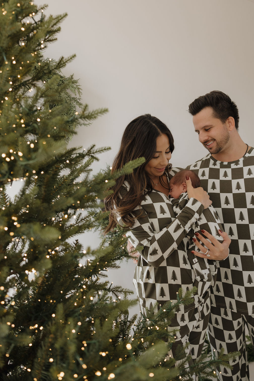 A smiling couple wears forever french baby Women's Bamboo Pajamas in the Christmas Tree Checkerboard print, holding a baby by a warmly lit, decorated Christmas tree for a cozy, festive scene.