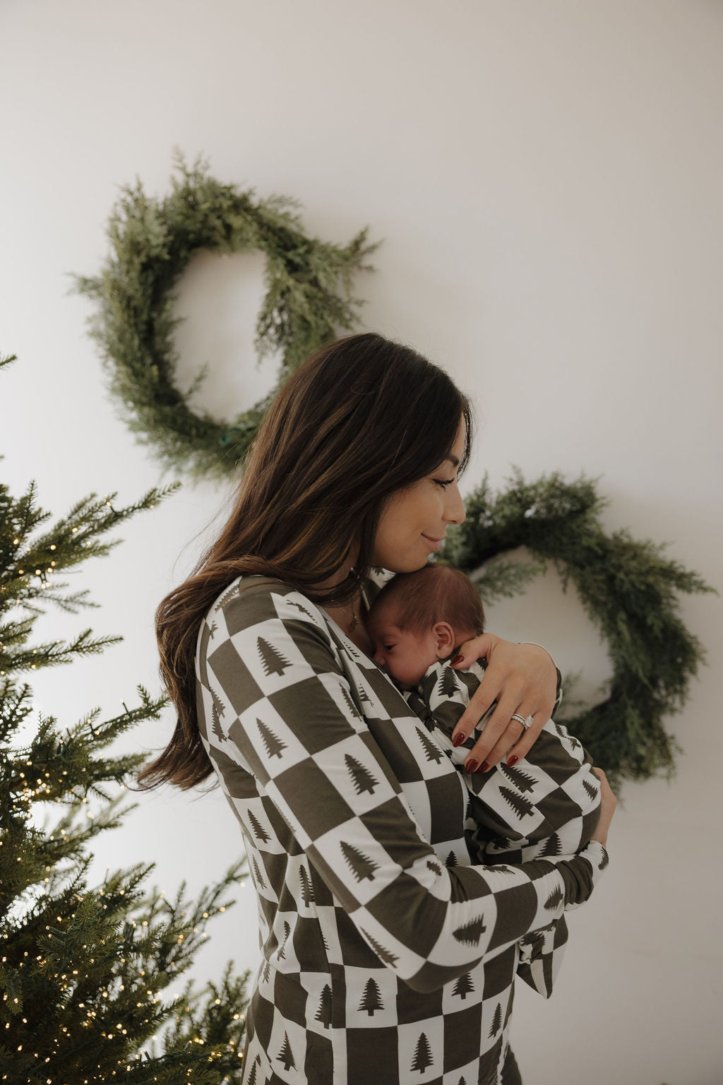 A woman and her newborn wear matching forever french baby Bamboo Zip Pajamas in the Christmas Tree Checkerboard print, standing by a lit Christmas tree and a white wall with green wreaths.