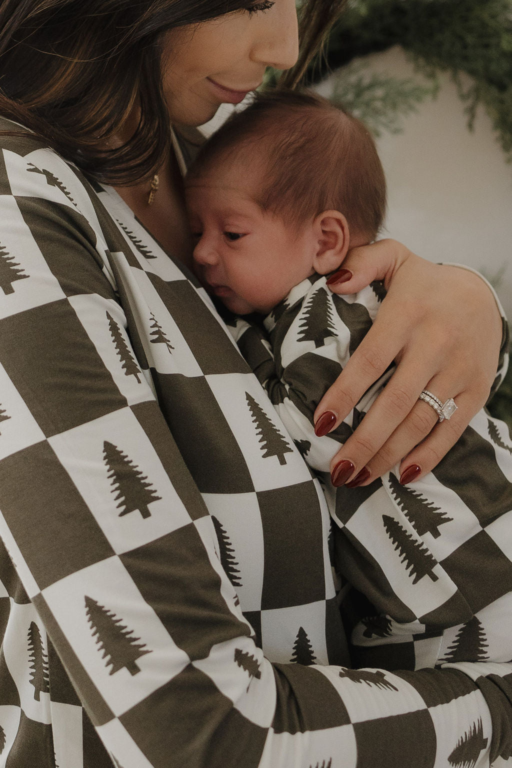 A woman with manicured nails and a wedding ring lovingly cradles her newborn in forever french baby's Bamboo Zip Pajamas | Christmas Tree Checkerboard, soft hypo-allergenic babywear, as greenery blurs in the background.