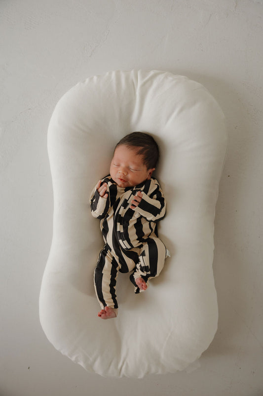 A newborn sleeps on a white, oval cushion, wearing forever french baby Bamboo Zip Pajamas in Midnight Stripe—a black and white striped onesie—hands relaxed by their face, feet bare, against a soft neutral background.