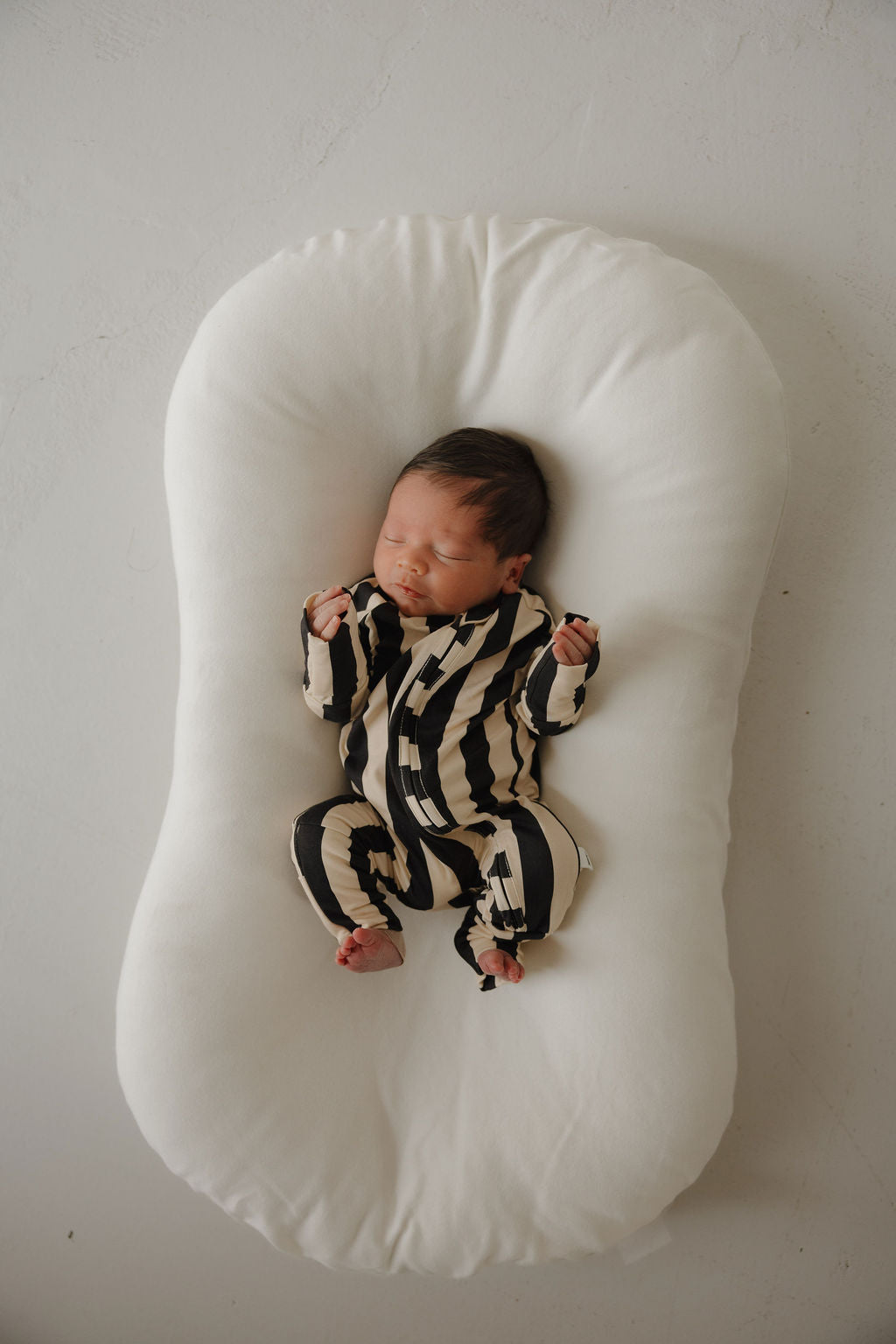 A newborn sleeps peacefully on a white cushion, dressed in forever french baby Bamboo Zip Pajamas | Midnight Stripe—breathable and hypoallergenic sleepwear—hands curled and eyes closed against a light background.