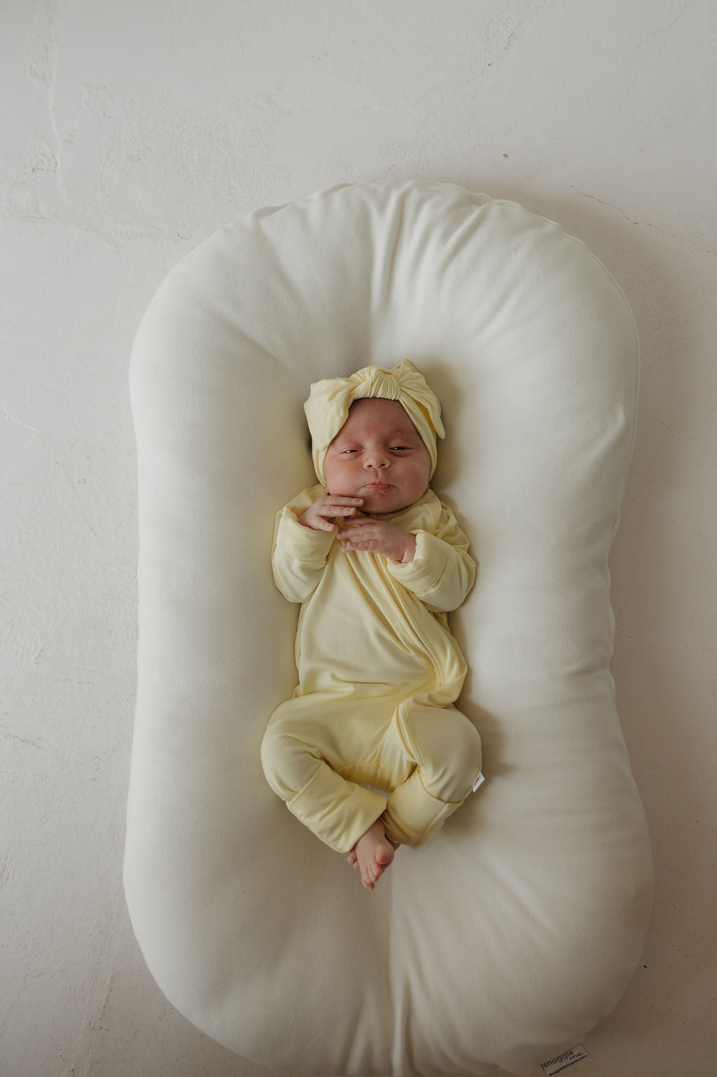 A baby wearing the forever french baby Bamboo Head Wrap | Sunny and a yellow outfit rests on a white cushioned pillow, eyes closed and hands near their face, against a light-colored textured background.