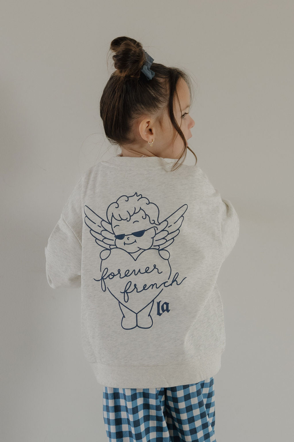 A young girl with brown hair in a bun wears the Child Sweatshirt by forever french baby, featuring a cartoon angel and "forever french" text on the back, paired with blue checkered pants, standing against a plain light gray background.