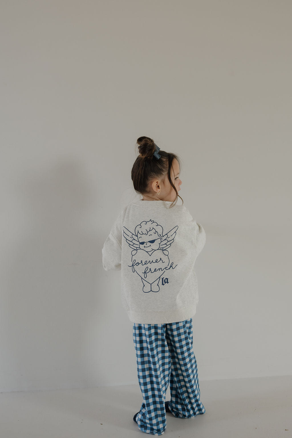 A young child with brown hair in a bun wears the forever french baby Child Sweatshirt | ff Los Angeles, featuring an angel graphic and "forever friend" on the back, paired with blue checkered pants against a plain light background.
