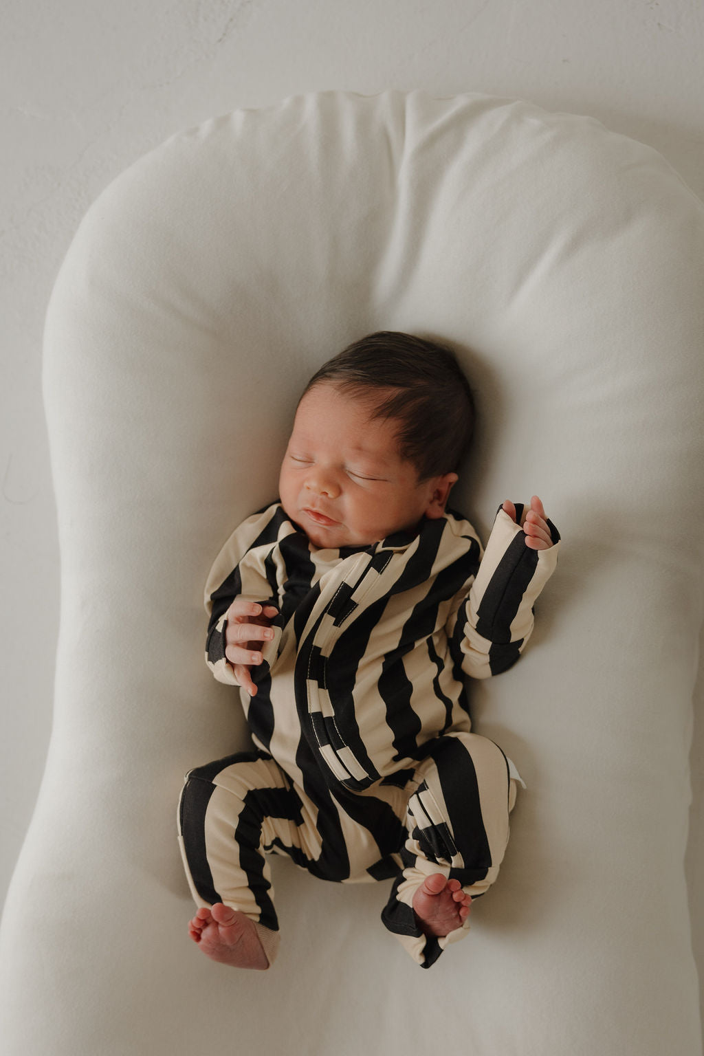 A newborn sleeps peacefully on a white cushioned surface, dressed in forever french baby’s Bamboo Zip Pajamas | Midnight Stripe. The breathable black and beige striped onesie keeps the baby comfy, with relaxed hands and bare feet.