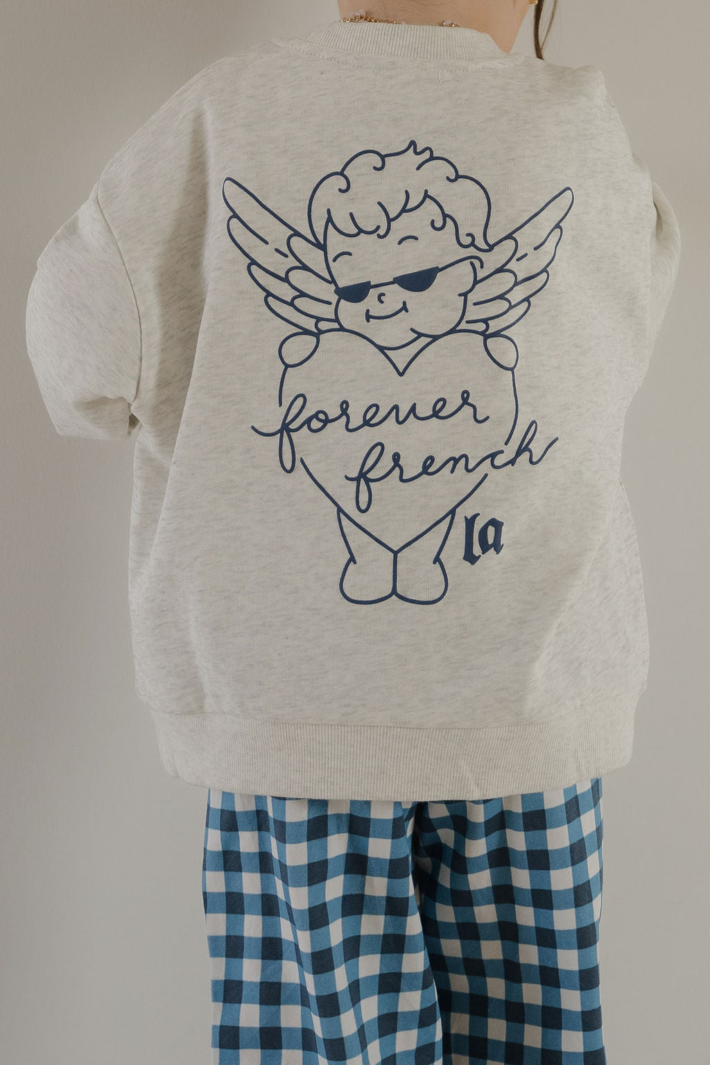 A child wears the forever french baby Child Sweatshirt | ff Los Angeles, featuring a winged cherub in sunglasses holding a "forever french" heart—perfect for matching family outfits.