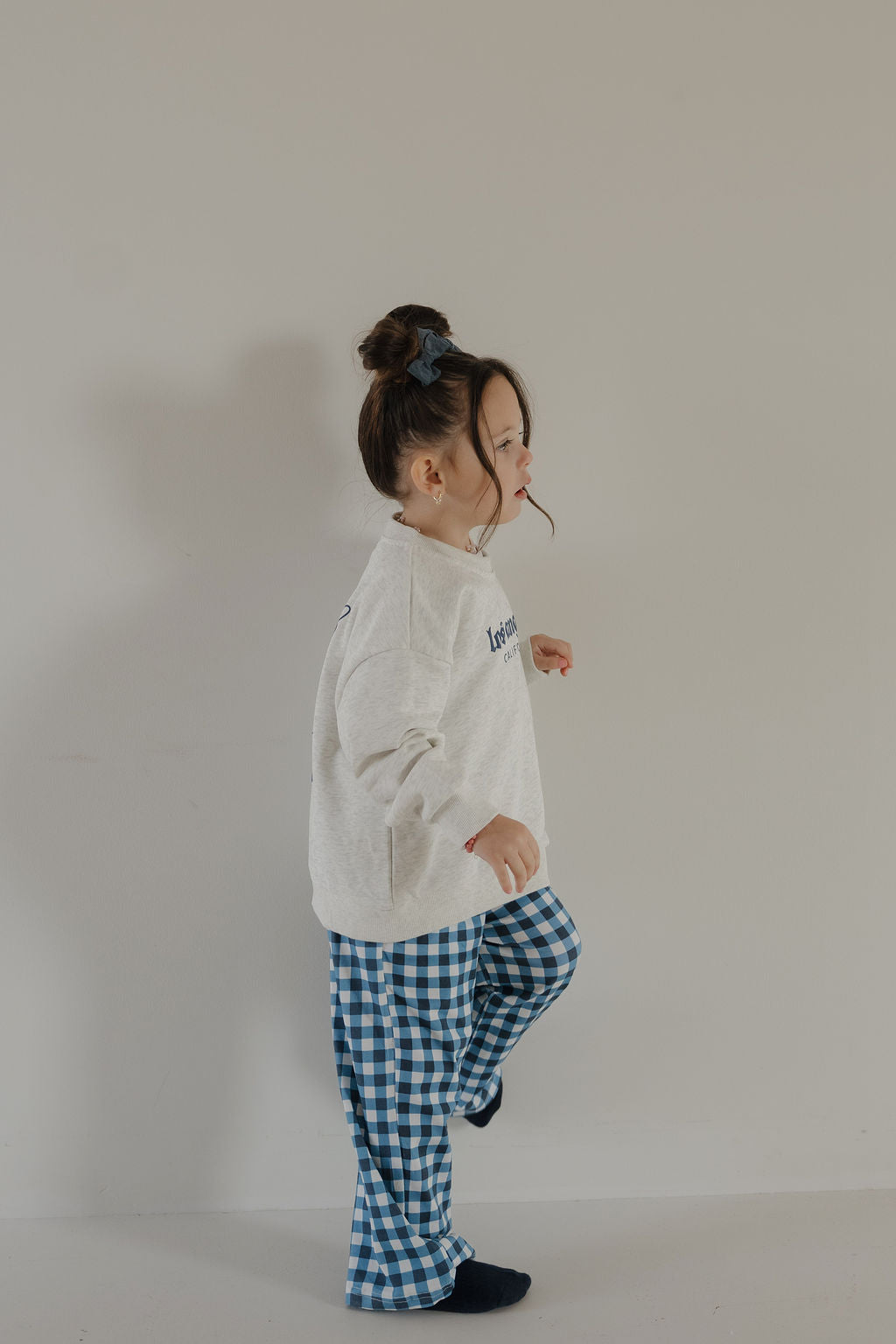 A young child with brown hair in a bun wears the forever french baby Child Sweatshirt | ff Los Angeles, blue and white checkered pants, and black socks while standing sideways with one leg lifted against a plain light-colored wall.
