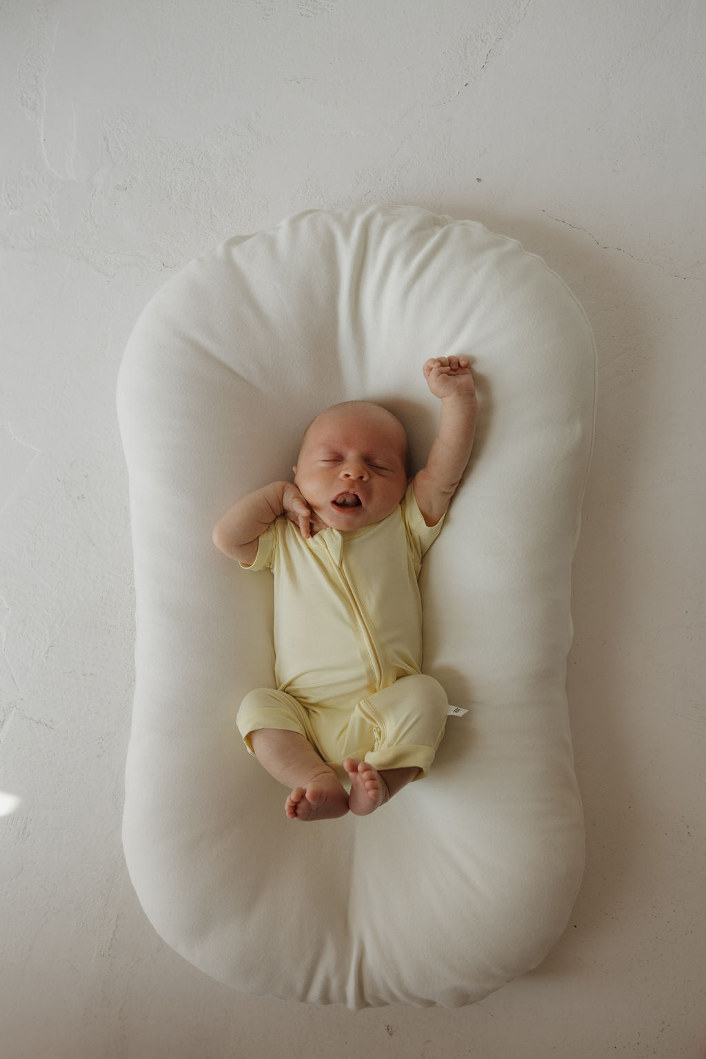 A newborn baby in the forever french baby Shortie Bamboo Zip | Sunny, a yellow onesie made from hypoallergenic bamboo fabric perfect for delicate skin, lies on a soft white cushion, stretching with one arm raised and eyes closed.