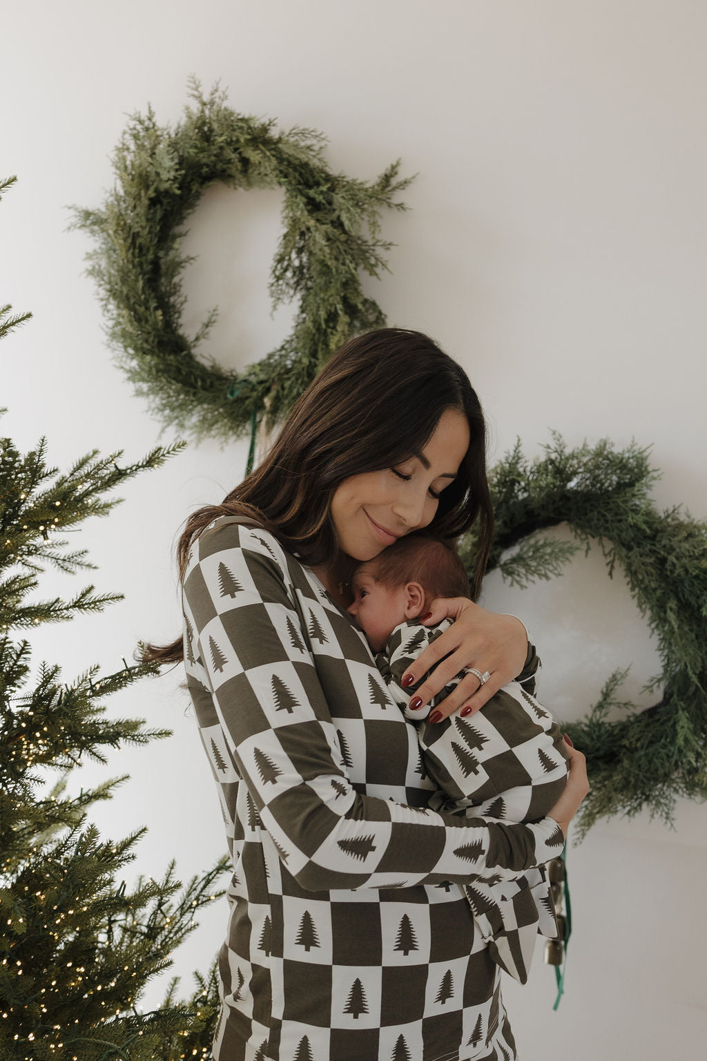 A woman hugs a baby, both in forever french baby Bamboo Zip Pajamas | Christmas Tree Checkerboard, standing by a Christmas tree with green wreaths on a white wall in the background.