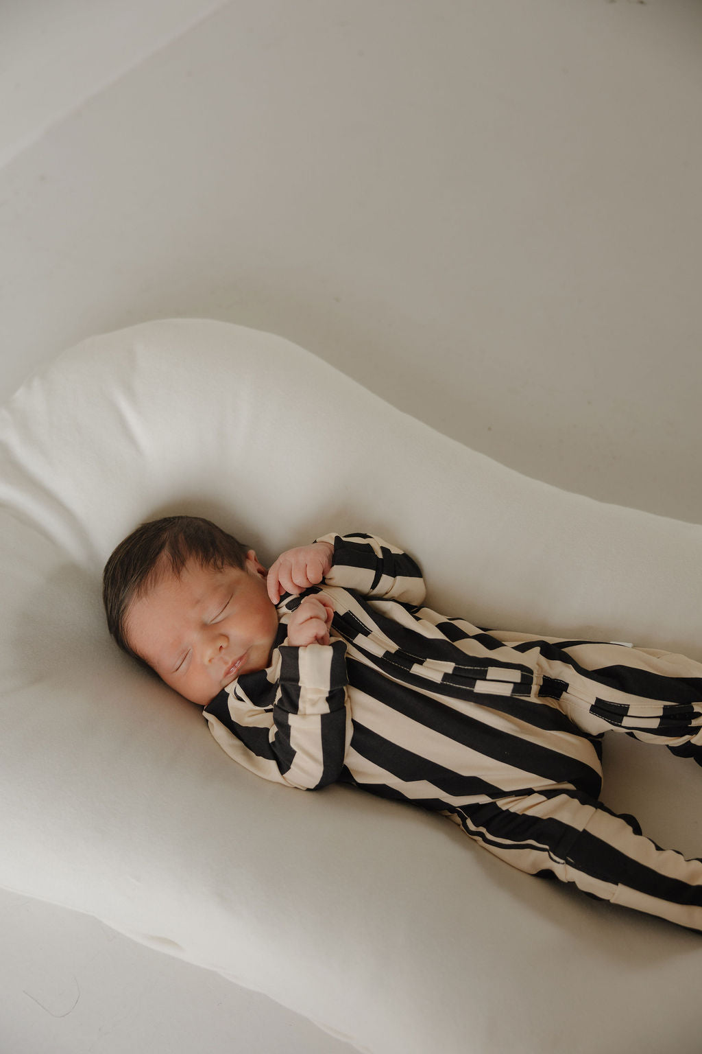A newborn with dark hair sleeps on a white cushion, wearing forever french baby Bamboo Zip Pajamas in Midnight Stripe. The black and beige striped sleepwear is breathable, with the baby's hands gently curled near their face.