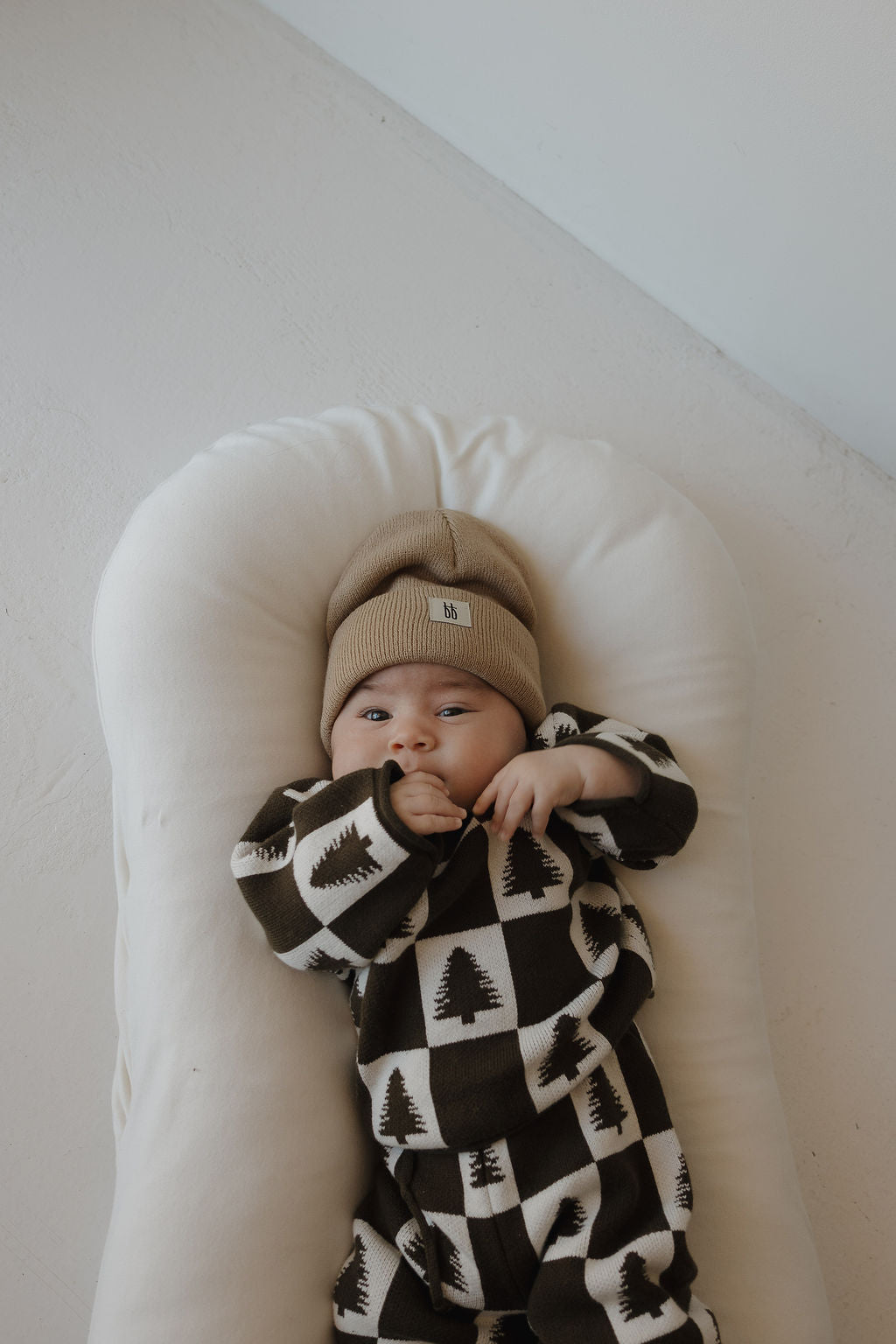 A baby wears the forever french baby Knit Pant Set | Christmas Tree Checkerboard— a cozy 100% cotton outfit with festive designs and a tan knit beanie, lying on a soft cushion and gazing up at the camera, hands near their mouth.