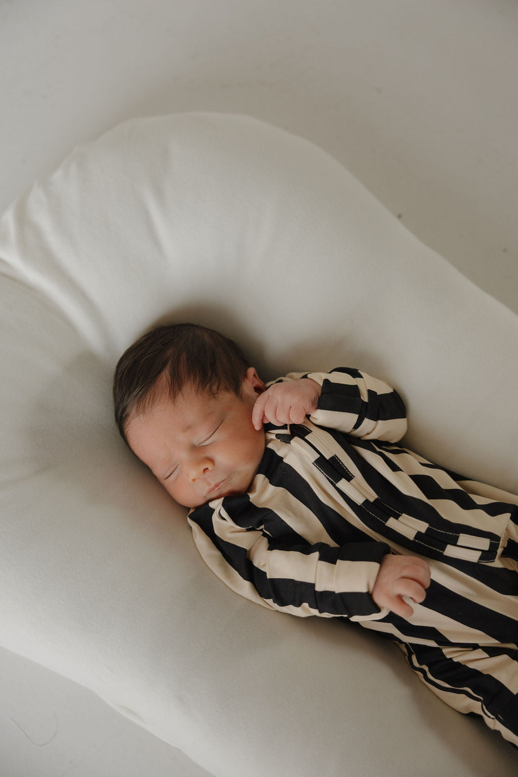 A newborn sleeps peacefully on a white pillow, dressed in forever french baby’s Bamboo Zip Pajamas | Midnight Stripe—comfortable, breathable black and beige striped sleepwear for all-night coziness.