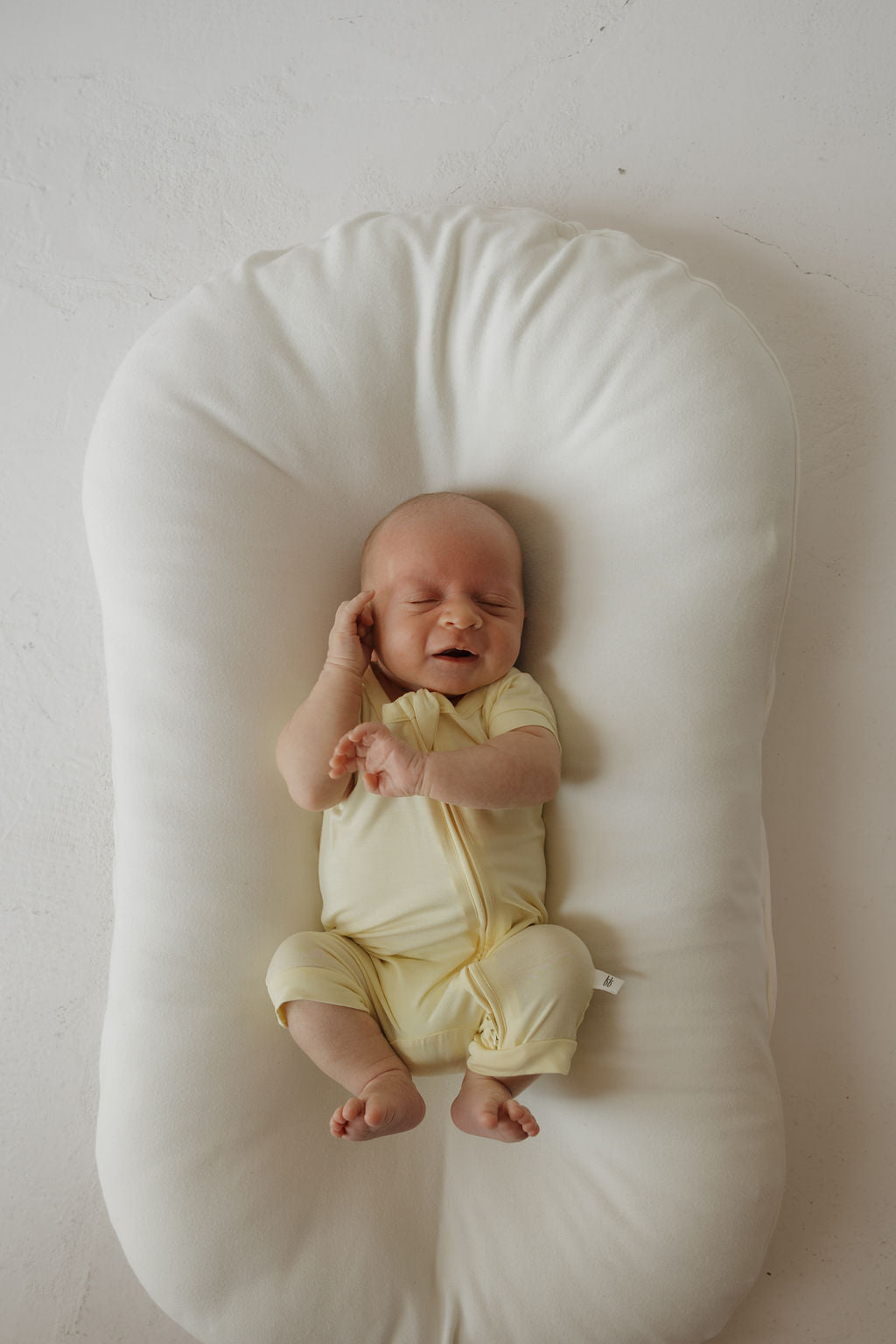 A newborn baby in the forever french baby Shortie Bamboo Zip | Sunny—a cheerful yellow bamboo romper—lies on a soft white cushion, eyes closed and smiling slightly, with one hand touching their head and legs gently bent, enjoying the gentle comfort of hypoallergenic clothing.