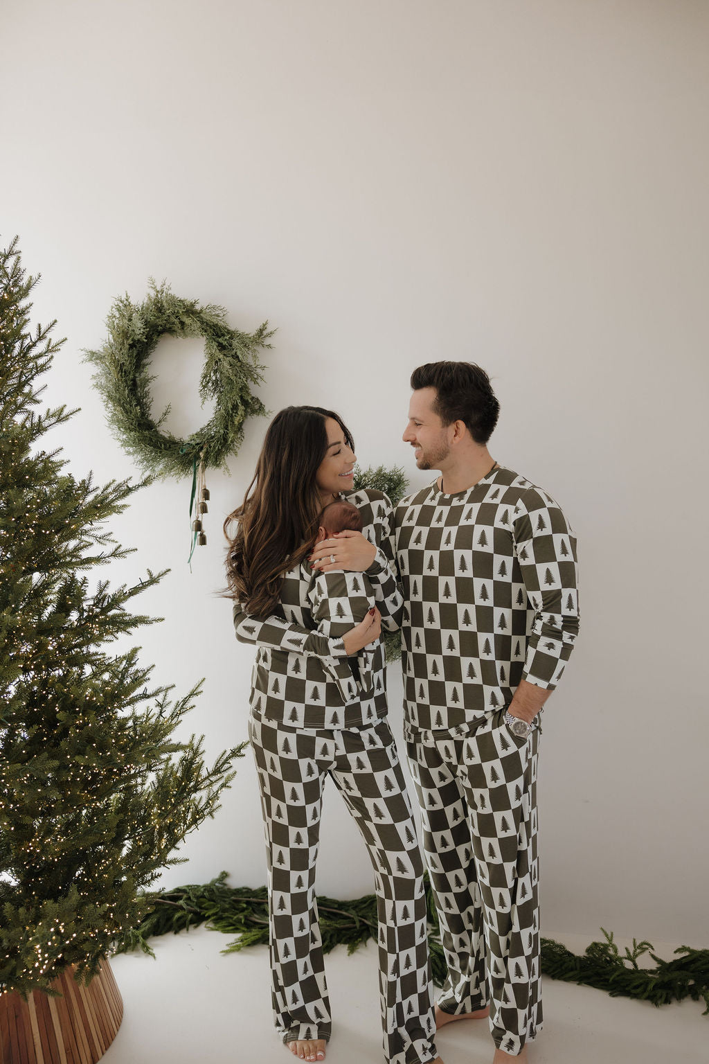 A couple smiles at each other beside a decorated tree, wearing forever french baby Men's Bamboo Pajamas in the Christmas Tree Checkerboard print—a festive, cozy, and hypo-allergenic choice for holiday comfort.