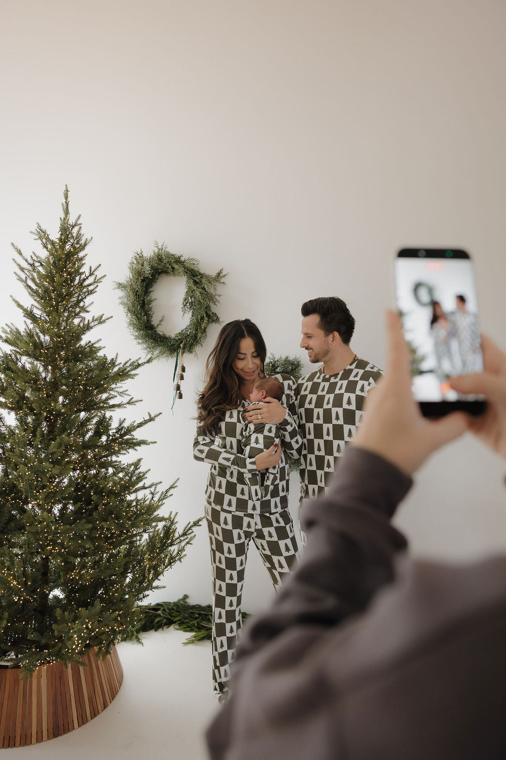 A family wears forever french baby Women's Bamboo Pajamas in Christmas Tree Checkerboard, posing with their baby by a festive tree and wreath as someone snaps a photo with a smartphone.