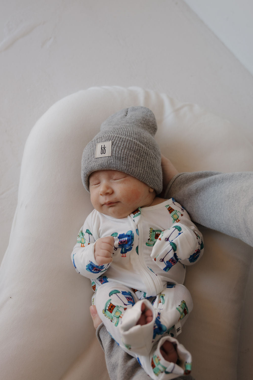A sleeping baby in forever french baby's Bamboo Zip Pajamas | Charli's Monsters lies on a soft white cushion, wearing a grey knit hat and supported gently by an adult’s hand, highlighting the charm of unique bamboo babywear.
