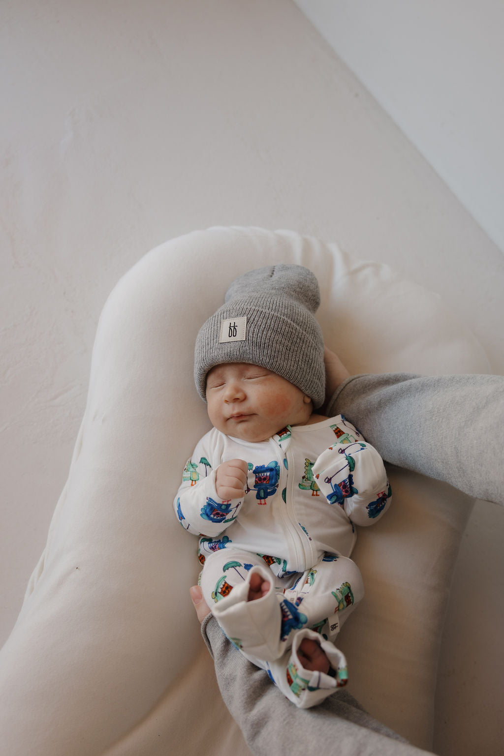 A newborn, eyes closed, lies on a white cushion wearing forever french baby's Bamboo Zip Pajamas | Charli's Monsters and a gray beanie, while an adult gently supports the baby's side.