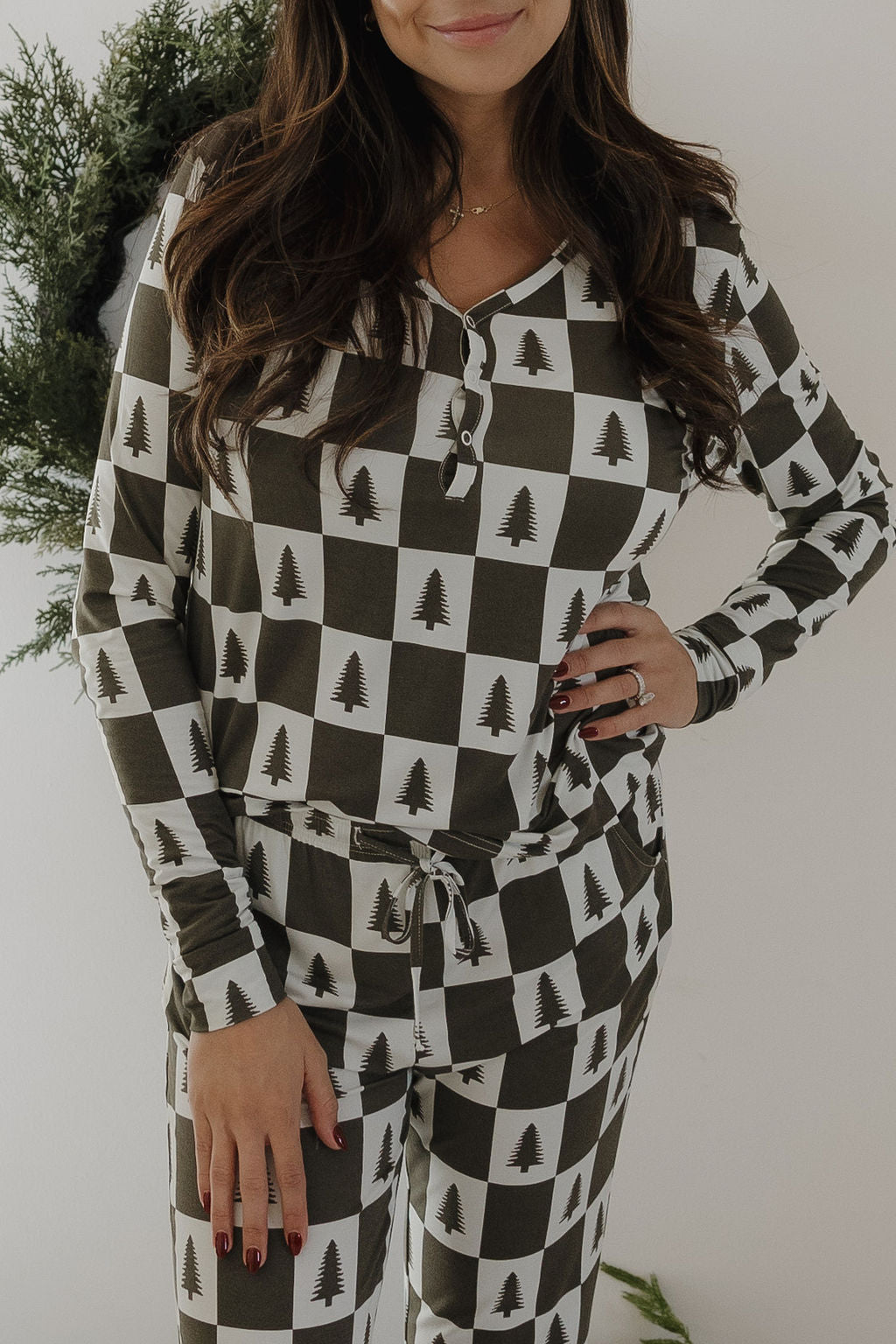 A woman stands confidently among greenery, wearing forever french baby Women's Bamboo Pajamas in a Christmas Tree Checkerboard pattern—black and white checks each with a festive tree, one hand on her hip.