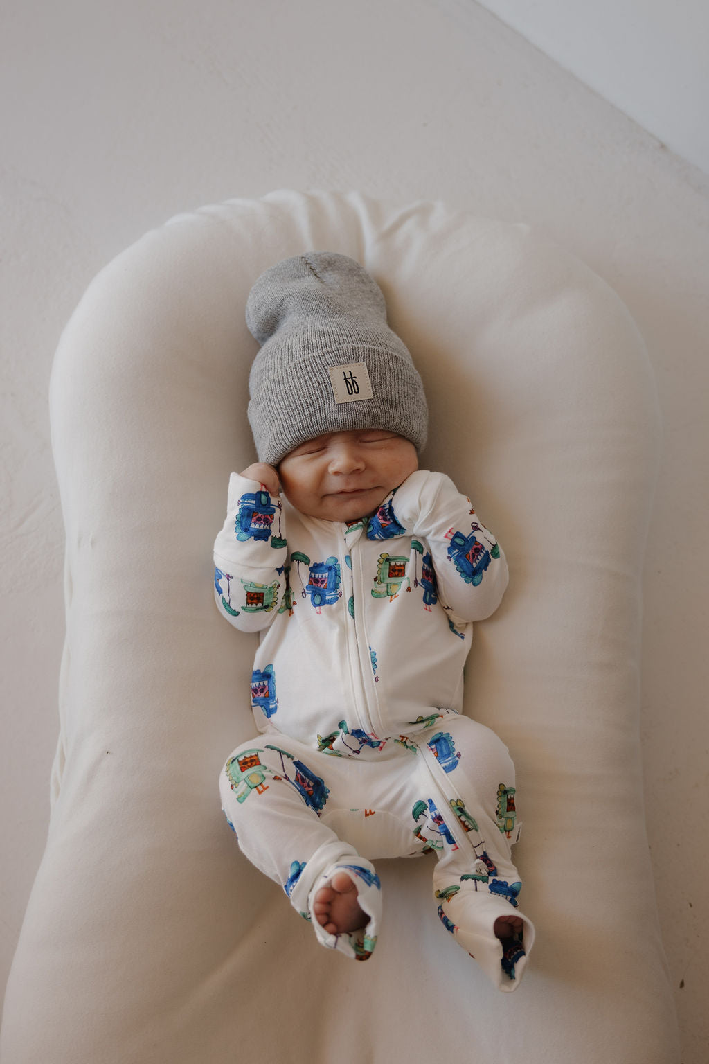 A smiling baby in forever french baby Bamboo Zip Pajamas | Charli's Monsters and a gray beanie lies on a soft white cushion, hands near their face.