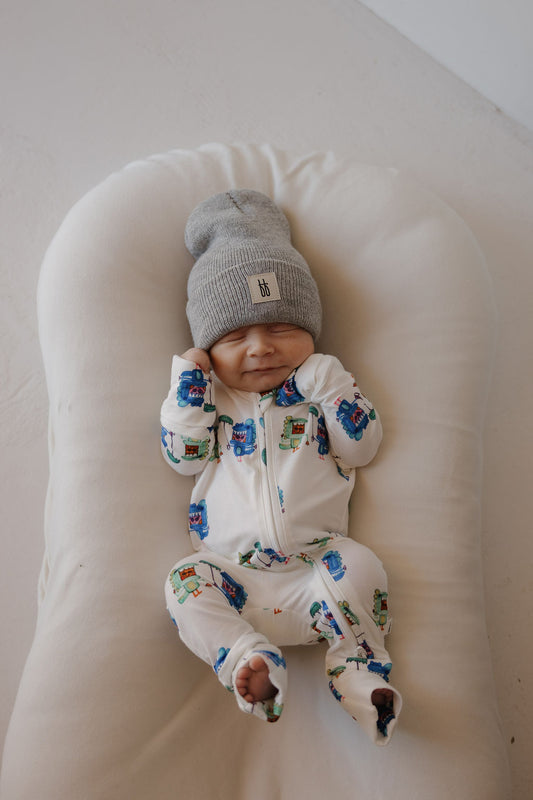 A smiling baby in forever french baby Bamboo Zip Pajamas | Charli's Monsters and a gray beanie lies on a soft white cushion, hands near their face.