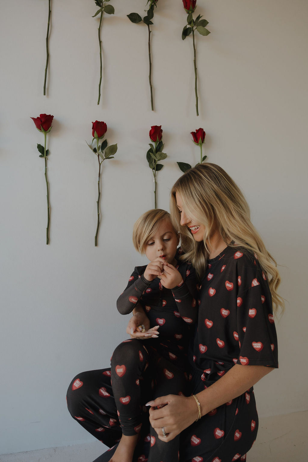 A woman and child, both in forever french baby Bamboo Two Piece Pajamas | Love Notes – Charcoal, featuring red heart prints, sit together by a wall of red roses, holding hands and enjoying their cozy bamboo outfits.