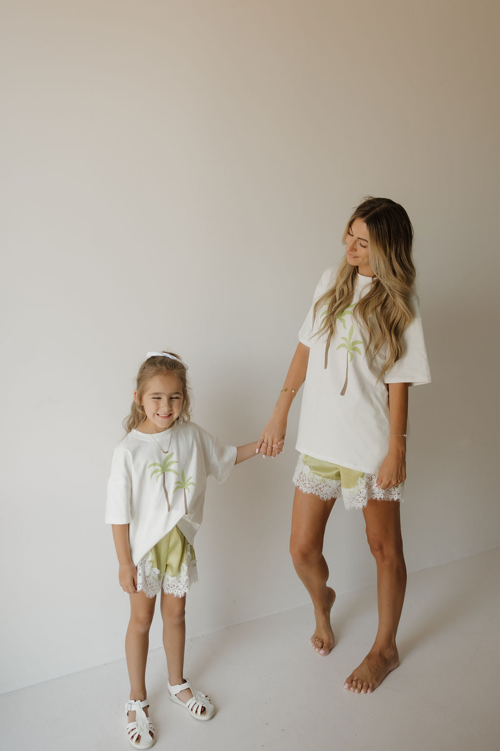 A woman and a young girl stand holding hands, both smiling against a plain beige wall in matching family outfits—each wearing the forever french baby Child Cotton Tee | Palm, crafted from 100% cotton with palm tree prints, paired with light green shorts featuring lace trim.