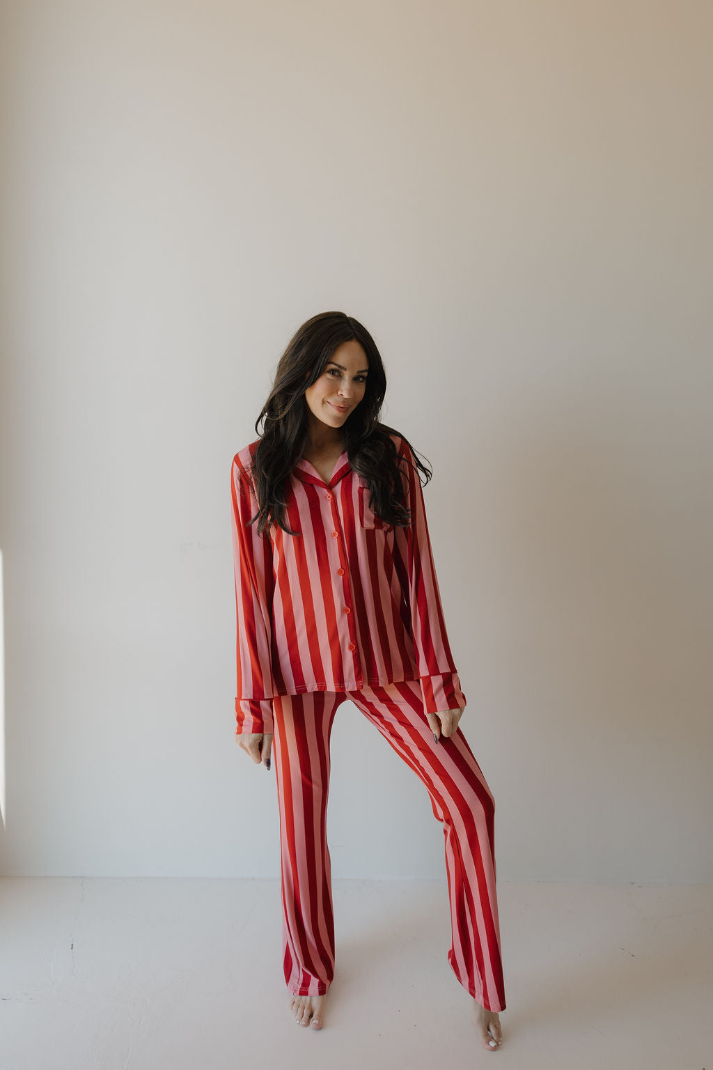 A woman with long dark hair stands barefoot against a plain light wall, wearing the Minty Cherries Women's Bamboo Button Up Lounge Set in Iconic Stripe, and smiles gently at the camera.