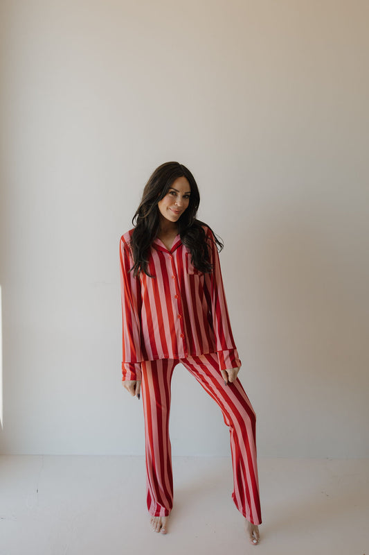 A woman with long dark hair stands barefoot against a plain light wall, wearing the Minty Cherries Women's Bamboo Button Up Lounge Set in Iconic Stripe, and smiles gently at the camera.