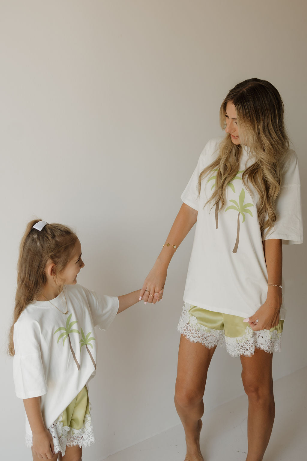 A woman and a young girl, both wearing matching palm tree shirts and forever french baby Girl’s Satin Shorts in Prickly Pear, hold hands and smile at each other against a plain light background.