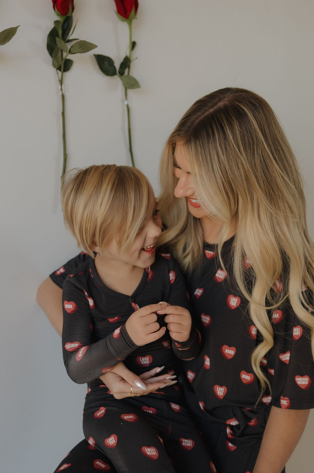 A woman and young child smile at each other in matching forever french baby Women's Boxy Tee Lounge Set | Love Notes - Charcoal, featuring red heart patterns. Two roses on the wall behind them add charm to their cozy moment.