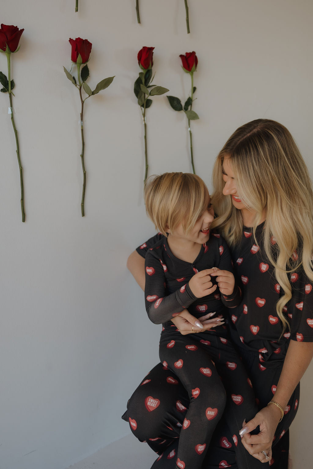 A woman and child smile together in matching forever french baby Women's Boxy Tee Lounge Sets in Love Notes - Charcoal, featuring red heart designs. Red roses are vertically attached to a white wall behind them.