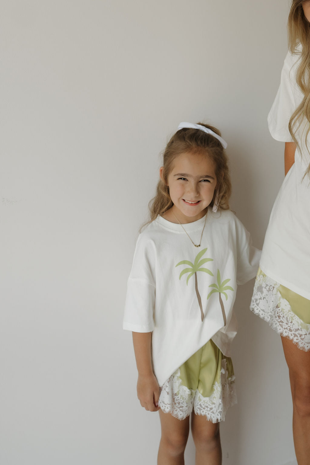 A young girl smiles, wearing the forever french baby Girl’s Satin Shorts in Prickly Pear—a green, lightweight style with lace trim—paired with a white palm tree shirt and a white hairband. She stands beside an adult in coordinating Forever French Baby outfits, both posed against a plain white wall.