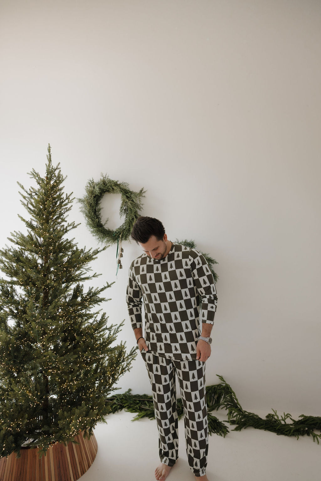 A person in forever french baby Men's Bamboo Pajamas | Christmas Tree Checkerboard stands barefoot by a decorated Christmas tree and green wreaths on a white wall, looking down as festive greenery adorns the floor.