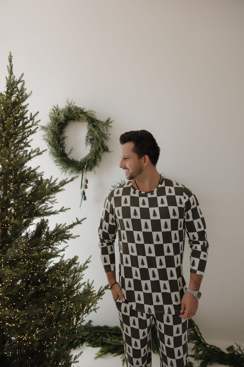 A man wearing forever french baby Men's Bamboo Pajamas | Christmas Tree Checkerboard smiles beside a decorated tree with a green wreath on the wall, adding festive charm to his cozy holiday look.
