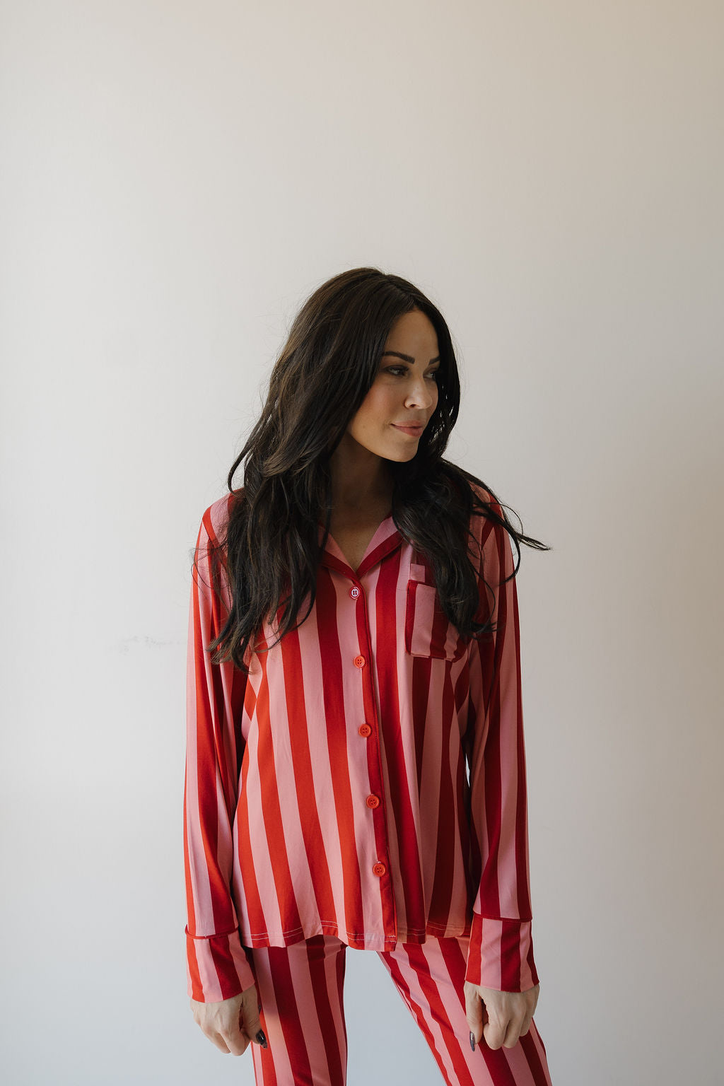 A woman with long dark hair models the Minty Cherries Women's Bamboo Button Up Lounge Set in Iconic Stripe, featuring red and pink vertical stripes, as she stands against a plain white background looking slightly left.